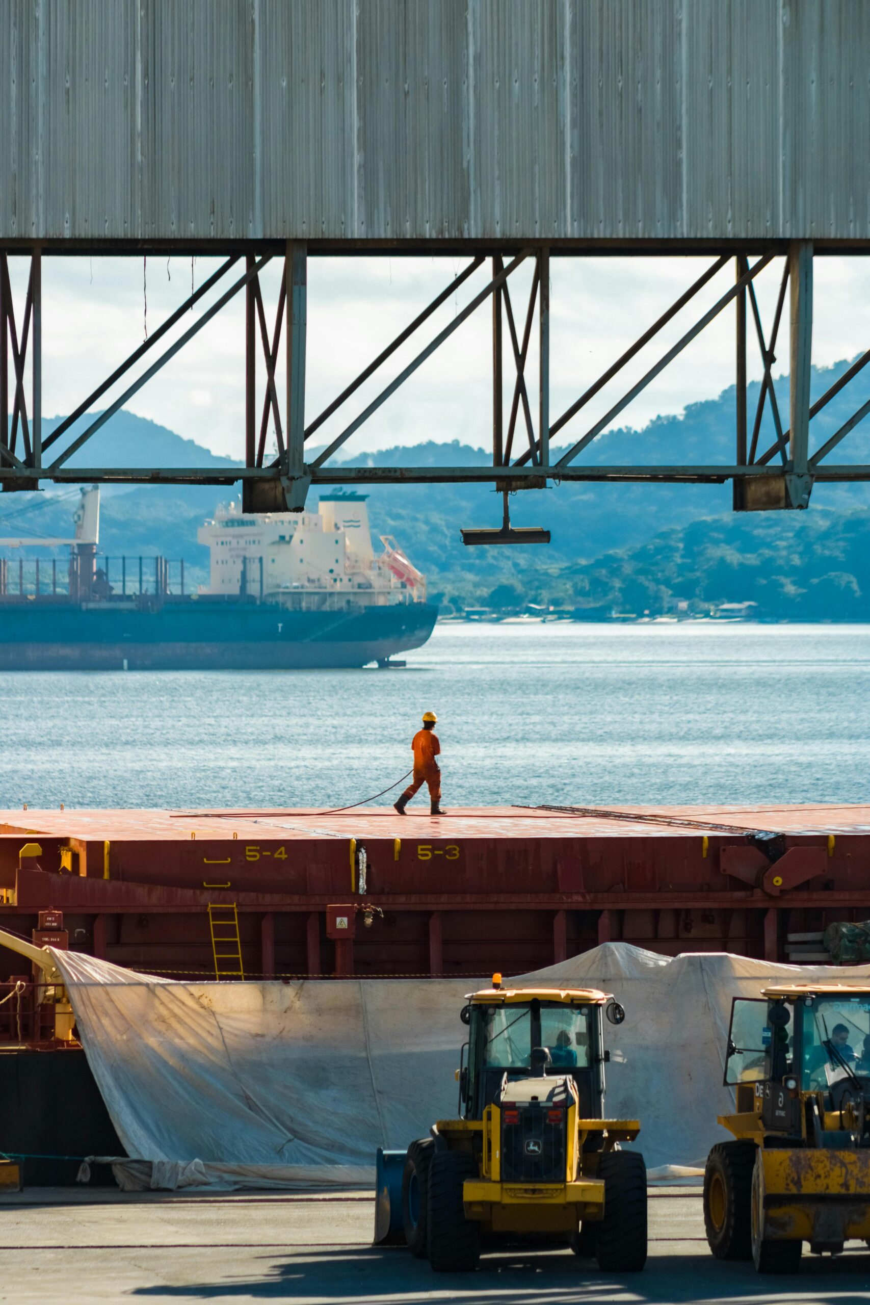 A dock worker in an orange uniform observes a cargo ship in an industrial harbor setting.